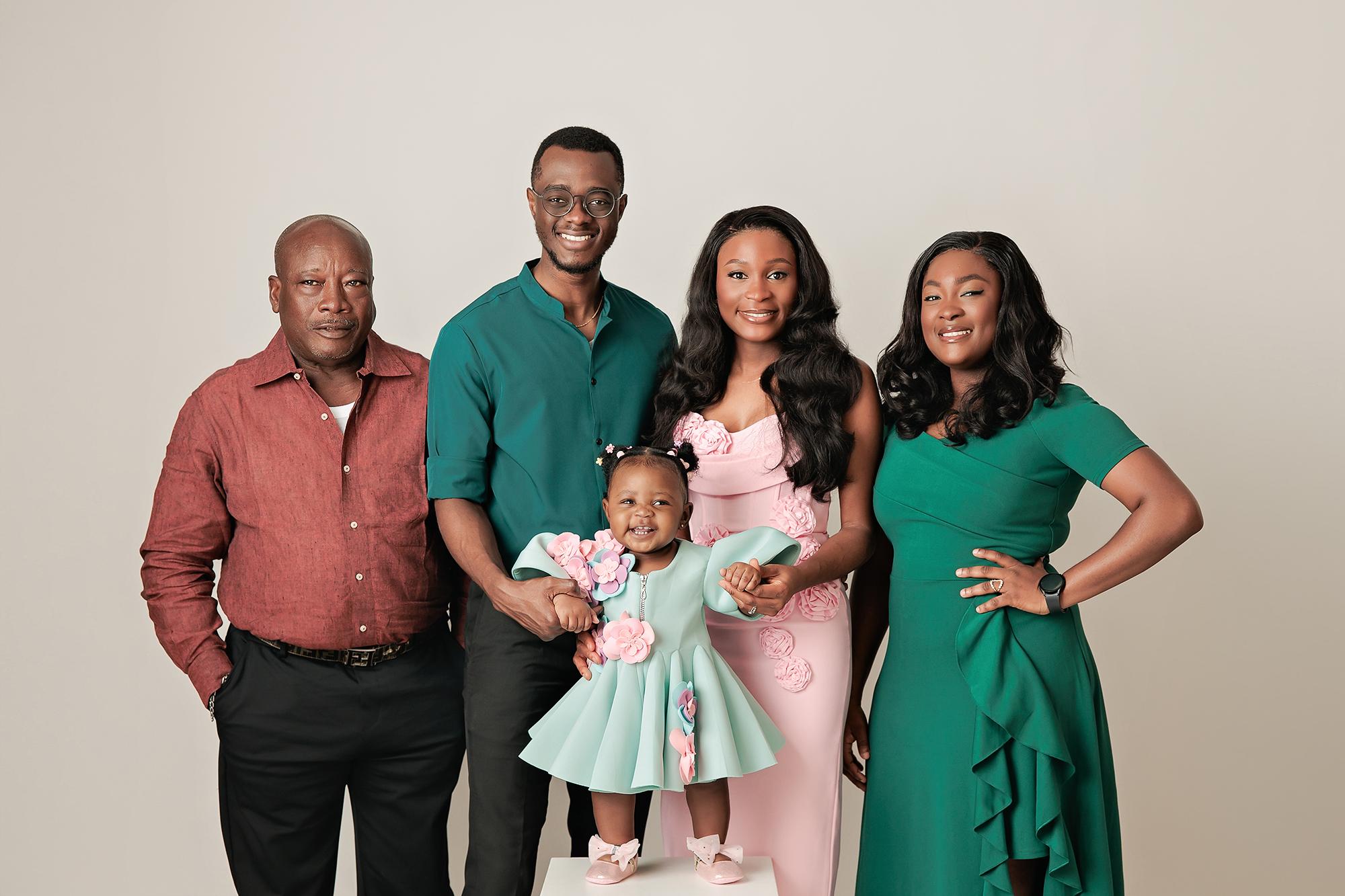 Extended family photography Calgary studio: Five adults and a baby girl in coordinated teal, pink, and red outfits, baby in a mint 3D floral dress on a white plinth, light grey background